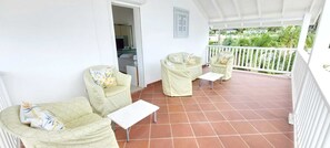Une terrasse couverte avec deux fauteuils vert clair dotés de coussins et une petite table, des balustrades blanches et une vue sur la verdure.