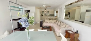 A modern living room with a glass dining table, a sofa, and a kitchen in the background. - 3BR Caribbean Home in Porters | Walk to Beach & Pool (Porters, Saint James)