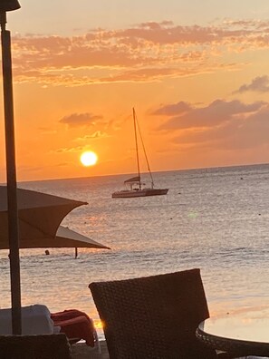 Une vue sur le coucher de soleil depuis un endroit situé au bord de la plage, avec un voilier à l’horizon et des chaises de plage au premier plan.