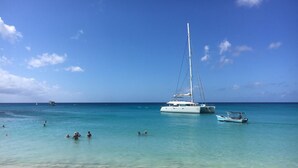 Ein Strand mit einem Segelboot, das in der Nähe der Küste vor Anker liegt, klarem türkisblauem Wasser und einigen darin schwimmenden Personen.
