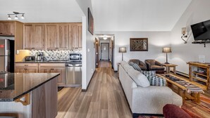 Een moderne keuken met houten kastjes, een granieten aanrecht en een ingebouwde oven. Een gezellige zithoek met een bank, fauteuils en een televisie is zichtbaar.