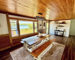 Interior em madeira com uma mesa grande e bancos, janelas amplas com vista para um lago e teto com vigas aparentes.