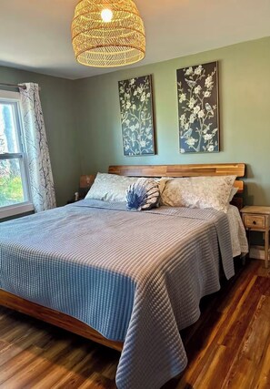 Quarto com uma cama grande, cabeceira de madeira e luminária pendente. Há duas pinturas fixadas na parede e uma janela com cortinas.