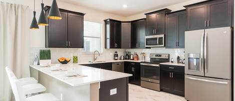 A modern kitchen with dark cabinets, stainless steel appliances, a white island with bar stools, and pendant lighting.