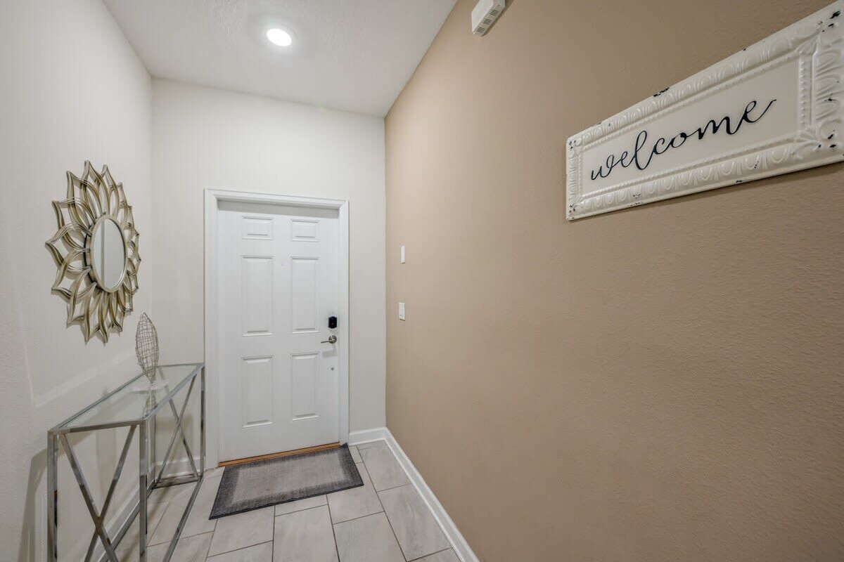 A hallway with a white door, a glass console table, a welcome sign, and a decorative mirror.