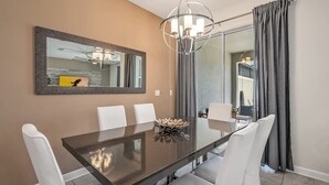 A dining area with a glass table, white chairs, a chandelier, a mirror, and grey curtains. - Magical Retreat, Spacious Villa 10 min From Disney (Kissimmee)