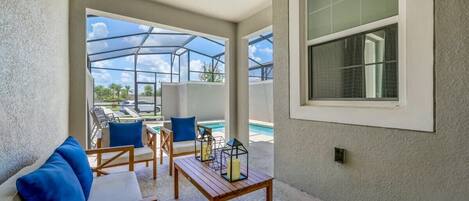 A covered outdoor seating area with a pool, wooden furniture, and a large window.