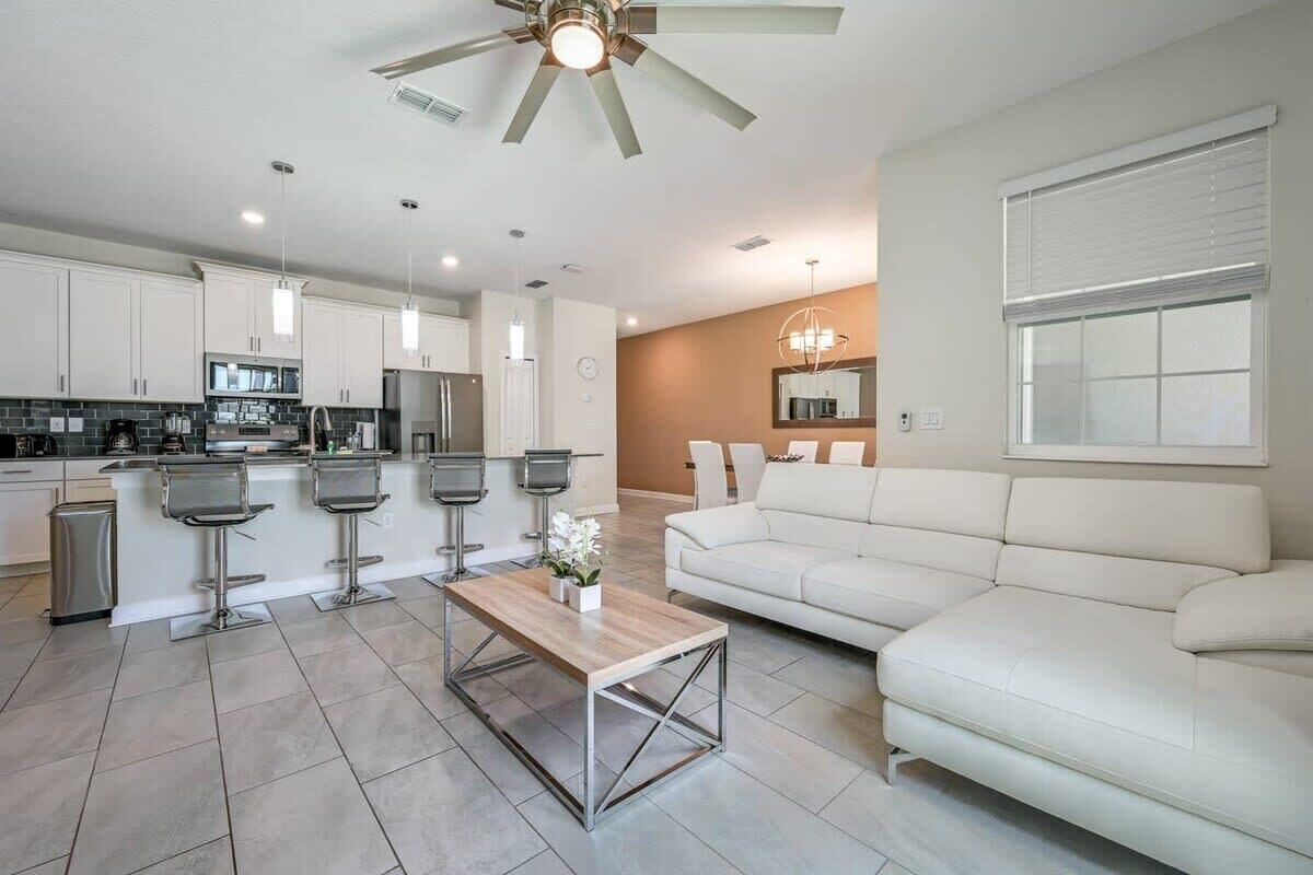 A modern kitchen with white cabinets, a central island with bar stools, a white sofa, and a ceiling fan.