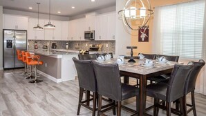 A modern kitchen with white cabinets, a stainless steel refrigerator, a granite countertop, and a dining area with a marble table and leather chairs. - Home 6BR - Prime Location (Kissimmee)