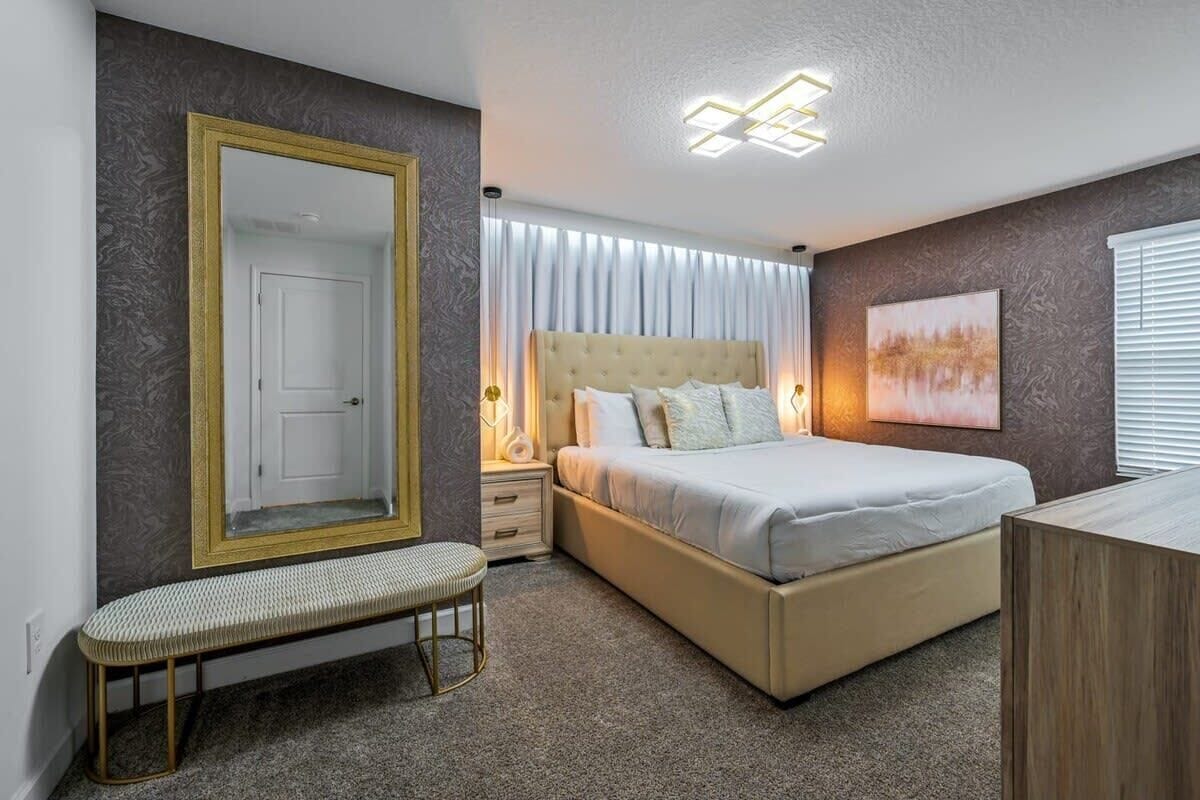 Une chambre à coucher avec un grand miroir, un lit doté d’une tête de lit capitonnée, un banc, une table de chevet et un luminaire fixé au mur.