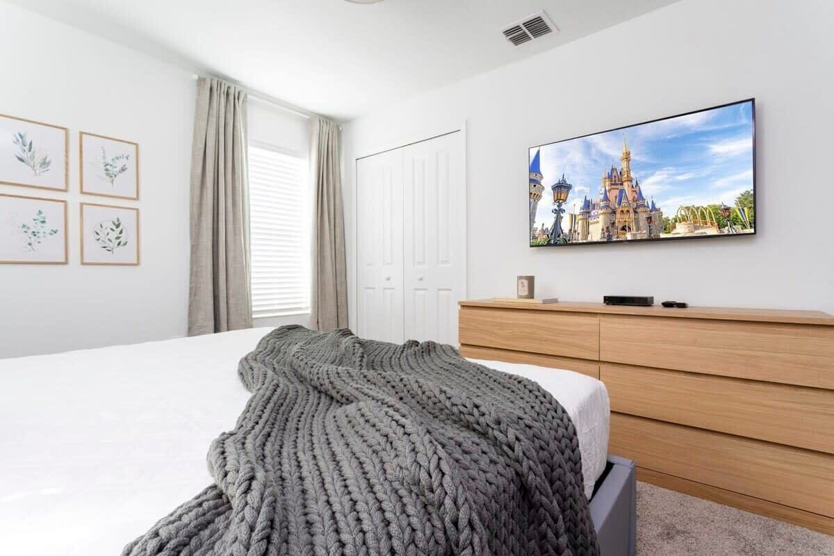Un dormitorio con cama, televisor en la pared, cómoda y ventana con cortinas.