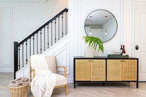 Uma escadaria com balaústres pretos, uma cadeira de vime com manta branca, um console de vime com espelho e uma planta.