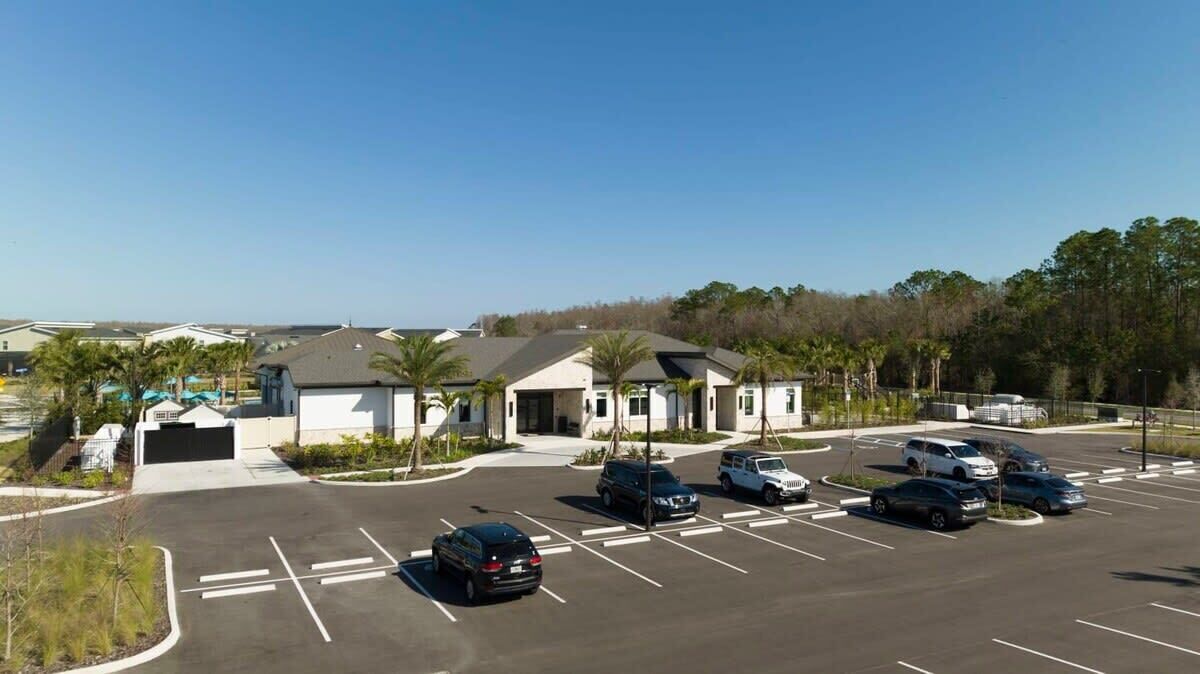 A parking lot with multiple cars, a building with a white facade and a dark roof, and palm trees in the background.