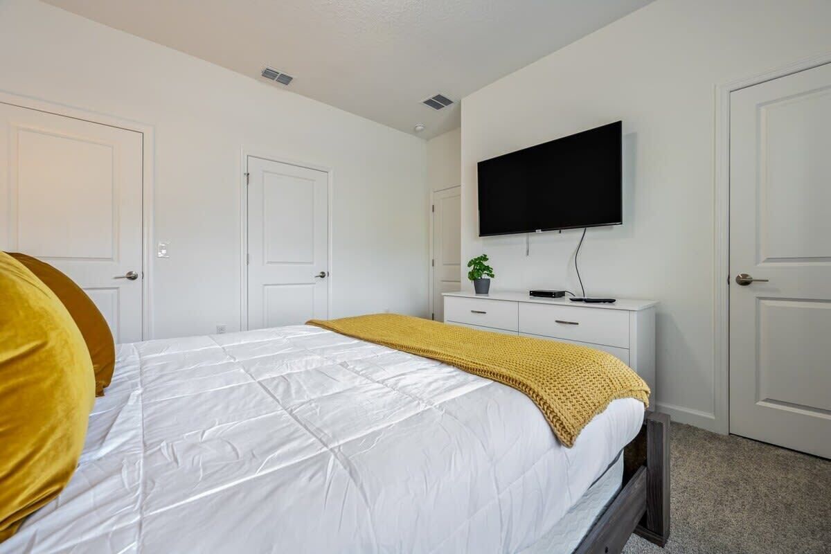A bedroom with a bed, a TV mounted on the wall, a bedside table with a plant, and a door.