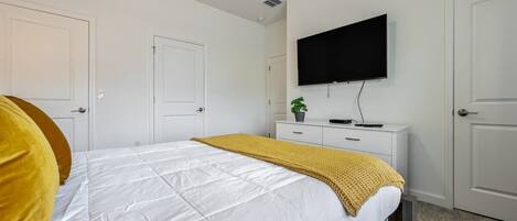 A bedroom with a bed, a TV mounted on the wall, a bedside table with a plant, and a door.