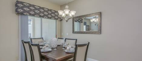 A dining area with a wooden table, six chairs, a chandelier, a large mirror, and patterned curtains.