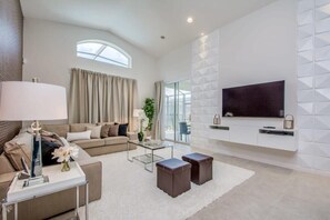 A modern living room with a sofa, coffee table, and ottomans, a flat-screen TV mounted on a white wall, and a large window with curtains. - Premium Comfort - Beautiful Home, Private Pool (Kissimmee)