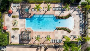 Ein Luftbild eines Resorts mit einem zentral gelegenen Swimmingpool, Liegestühlen und Palmen.
