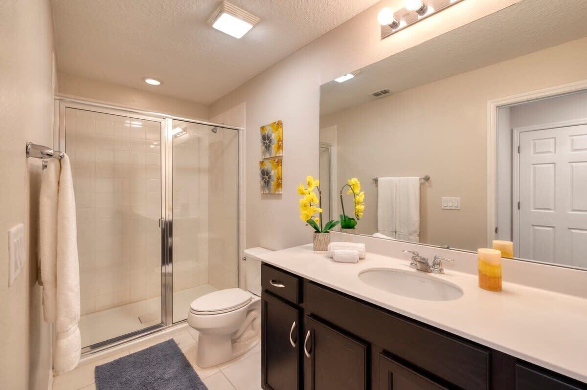Une salle de bain avec une douche à parois de verre, des toilettes, un lavabo avec un miroir et un vase rempli de fleurs jaunes.