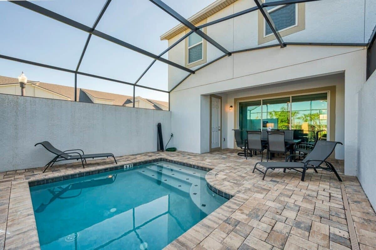 Una piscina con cerramiento de vidrio, sillas de descanso y un patio con mesa y sillas.