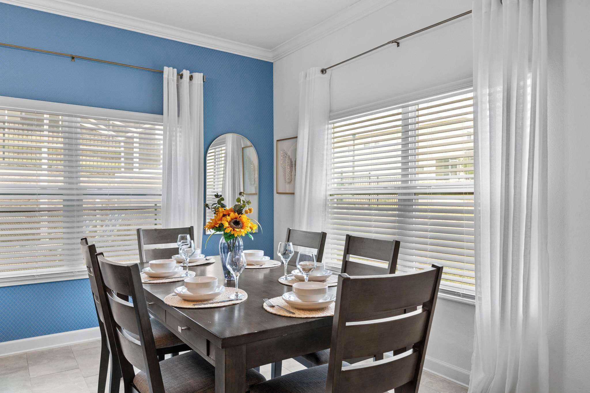 Une salle à manger avec une table en bois foncé, dressée pour quatre personnes, des rideaux blancs et un mur d’accent bleu.
