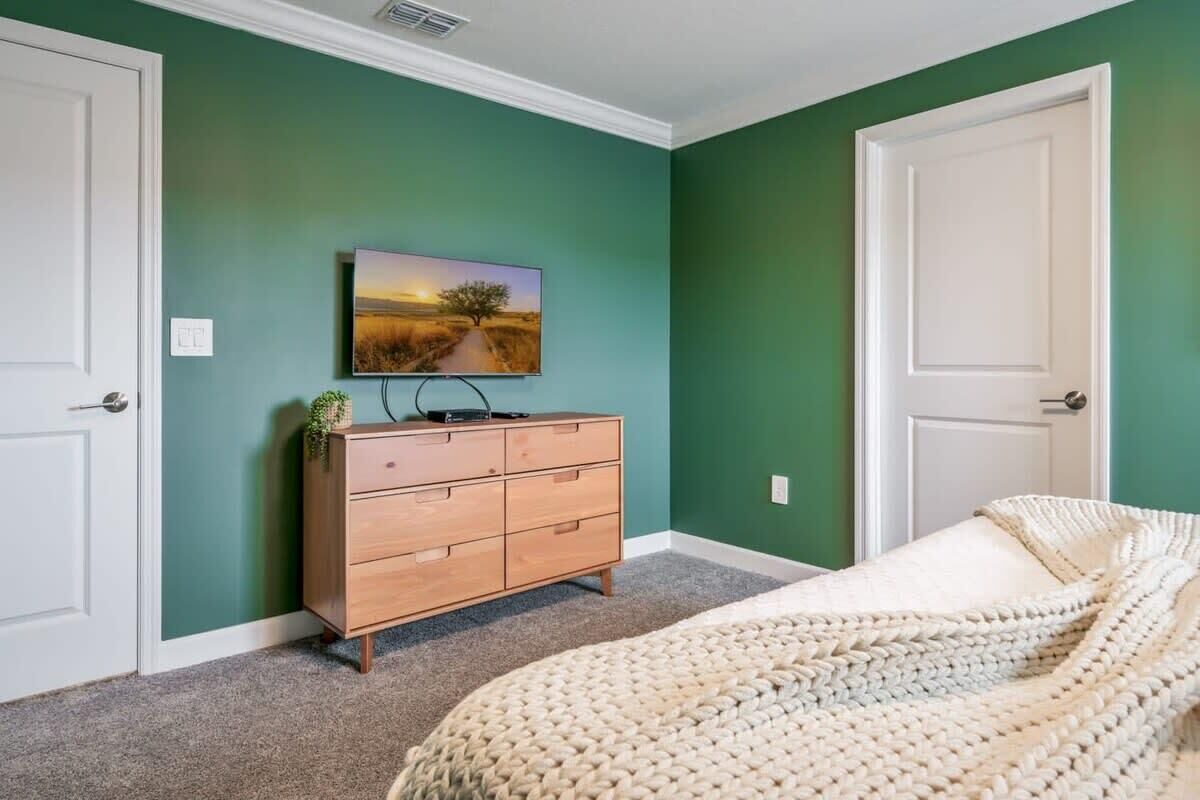 Un dormitorio con cama, cómoda, televisor en la pared y dos puertas.