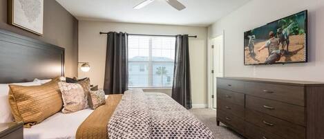 A hotel room with a bed, a dresser, a ceiling fan, a window with curtains, and a wall-mounted TV.