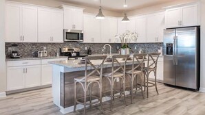 A modern kitchen with white cabinets, a stainless steel refrigerator, a granite countertop, and a central island with bar stools. - Chic Luxury Haven - Unforgettable Retreat (Kissimmee)
