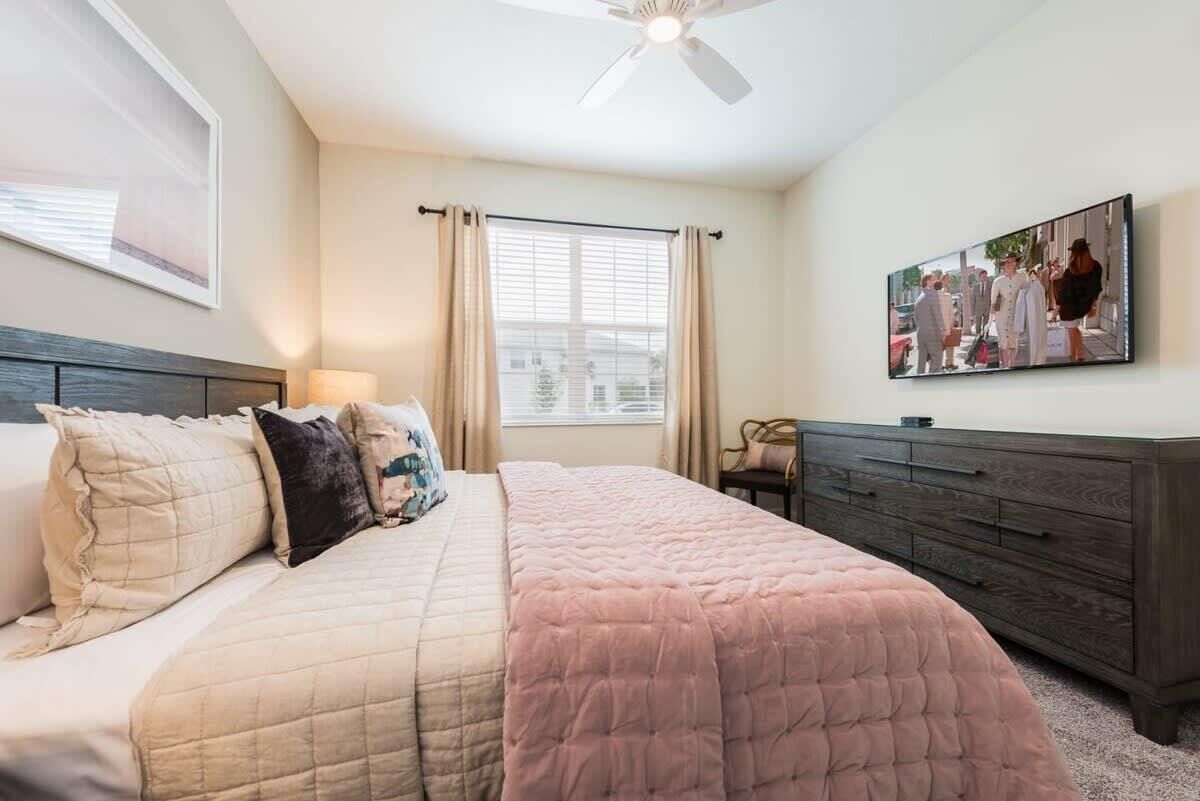 A bedroom with a bed, a dresser, a ceiling fan, a window with curtains, and a wall-mounted TV.