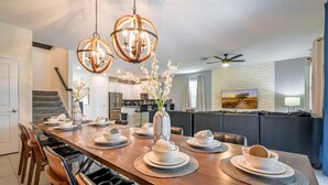 A dining area with a long wooden table set for a meal, a kitchen in the background, and a modern ceiling fan. - Family Paradise, Space, Themed, Perfect Location (Kissimmee)
