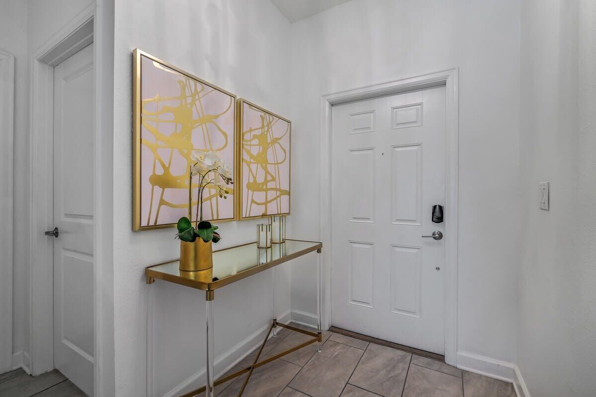 A hallway with a white door, a glass console table with a gold frame, and two gold-framed abstract paintings.