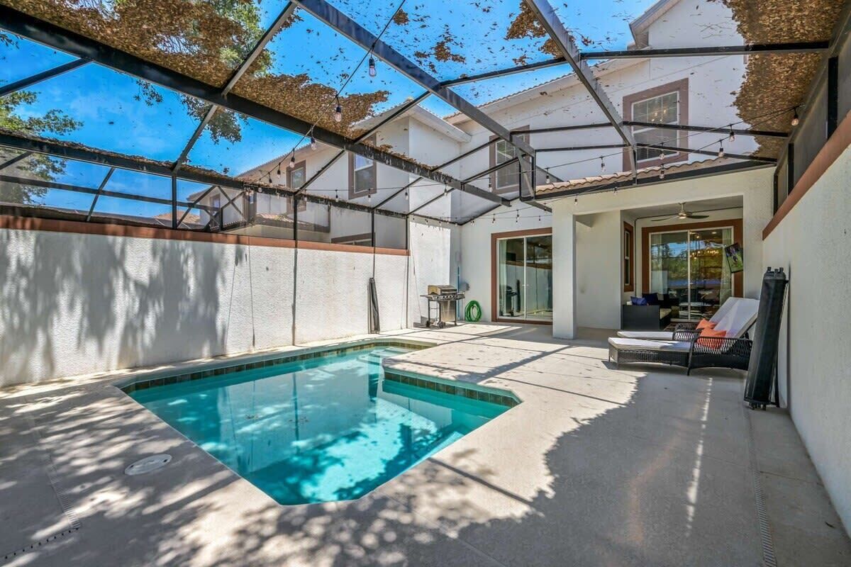A swimming pool with a glass enclosure, a covered patio area, and a seating area with a table and chairs.