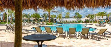 A poolside area with lounge chairs and a round table, surrounded by palm trees and a thatched roof structure.