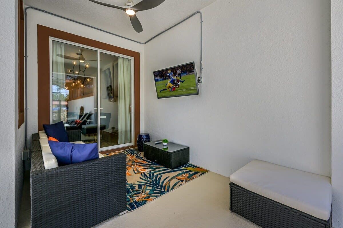 A balcony with wicker furniture, a TV mounted on the wall, and a glass door leading to an indoor area.