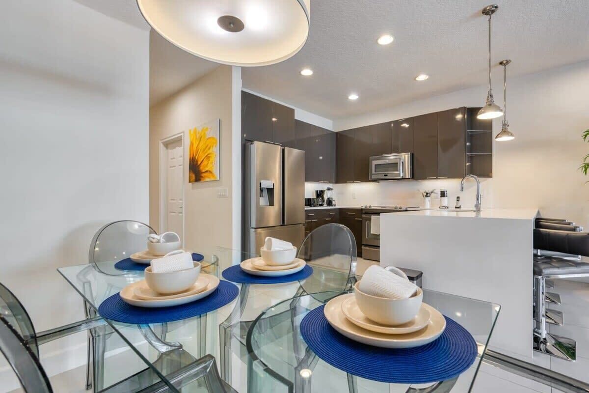 A modern kitchen with stainless steel appliances, a dining area with glass tables and blue placemats, and a central island with a sink.