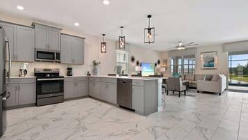 A modern kitchen with grey cabinets, a built-in microwave, and a dishwasher. There is a dining area with a white sofa and a TV.