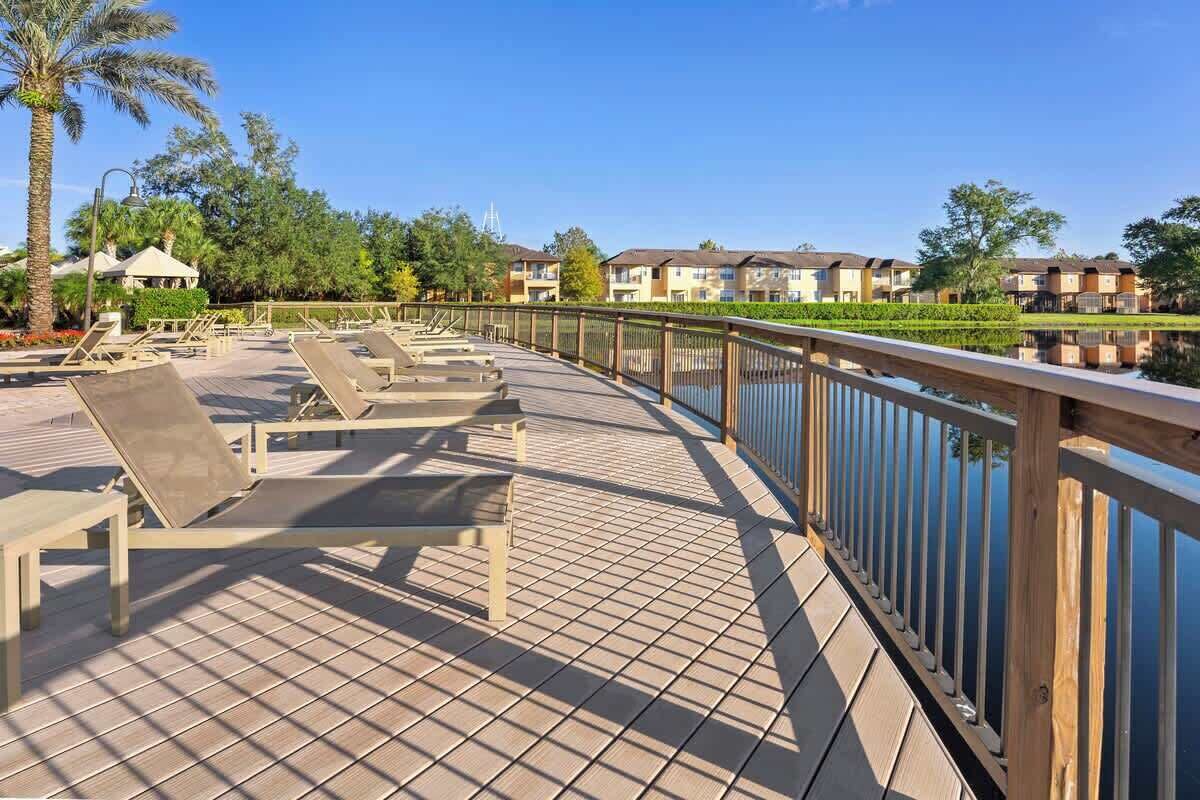 A wooden deck with sun loungers overlooking a calm body of water, surrounded by greenery and residential buildings.
