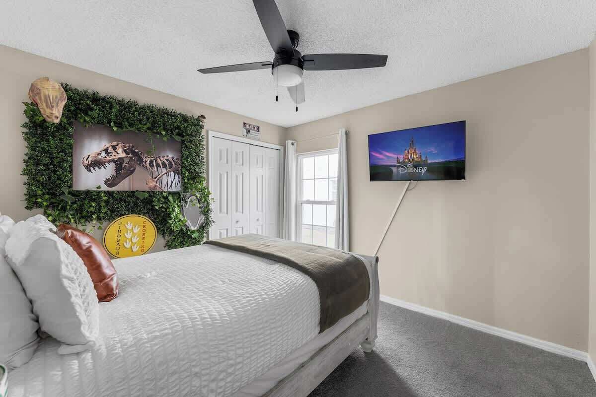 Ein Schlafzimmer mit einem Bett, einem Deckenventilator, einem Fernseher, einer Tür und einer Wand, die mit einem Dinosaurierskelett und Grünpflanzen verziert ist.
