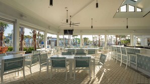 Un restaurante al aire libre con mesas y sillas blancas, una zona de bar y un ventilador de techo.