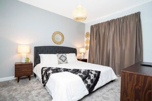 Un dormitorio con una cama grande, dos mesitas de noche, una lámpara colgante, un espejo redondo y un ventanal con cortinas.