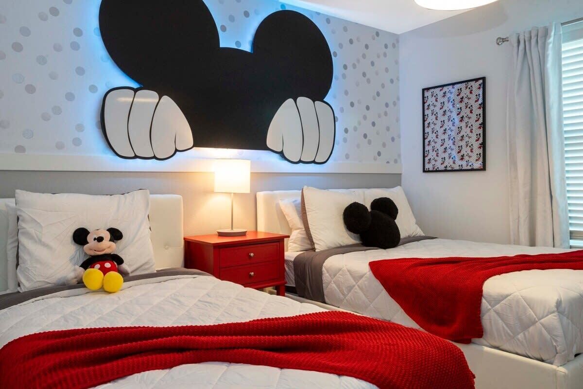 A hotel room with two beds, a Mickey Mouse pillow, a large Mickey Mouse wall decoration, a red blanket, and a nightstand with a lamp.