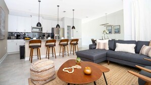 A modern kitchen with white cabinets, a dining area with a round wooden table, and a dark grey sofa in a living room. - Comfy Themed Home, Private Pool, Prime Location (Davenport)