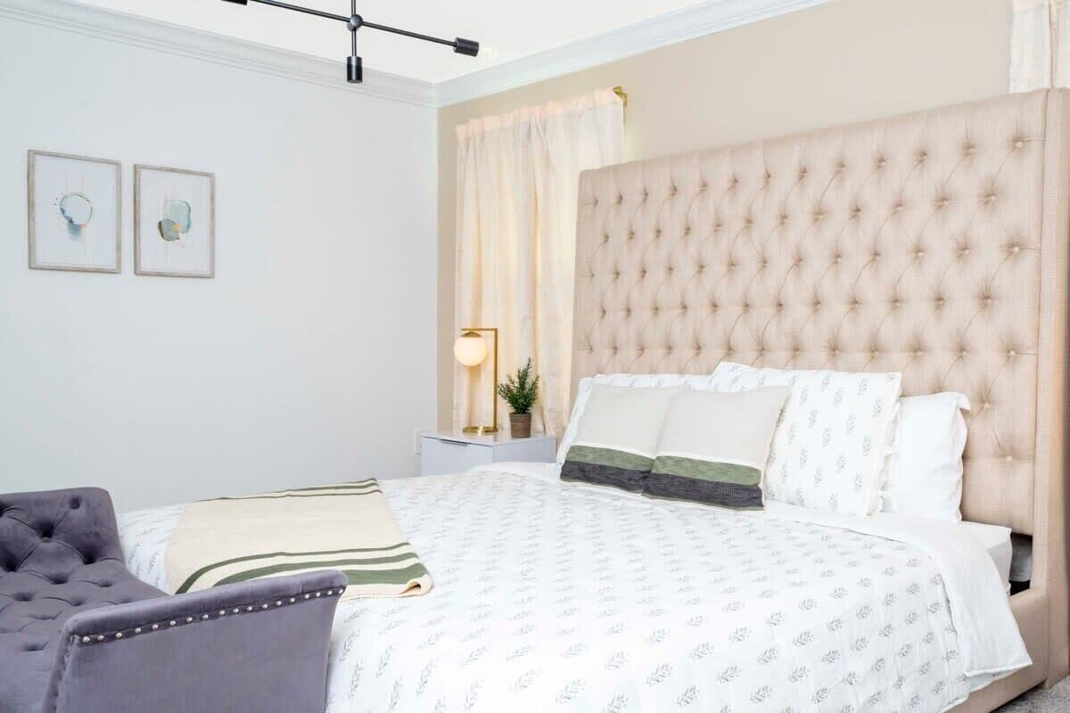 A neatly arranged bedroom with a tufted headboard, a bed with patterned bedding, a grey armchair, and two framed artworks on the wall.