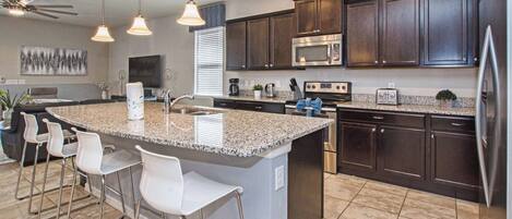 A modern kitchen with dark wood cabinets, a granite countertop, stainless steel appliances, and a bar area with white stools.