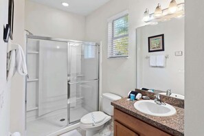 A modern bathroom with a glass-enclosed shower, a white toilet, a sink with a mirror, and a window with blinds. - Lake View and Private Pool 4BR at Bella Vida (Kissimmee)