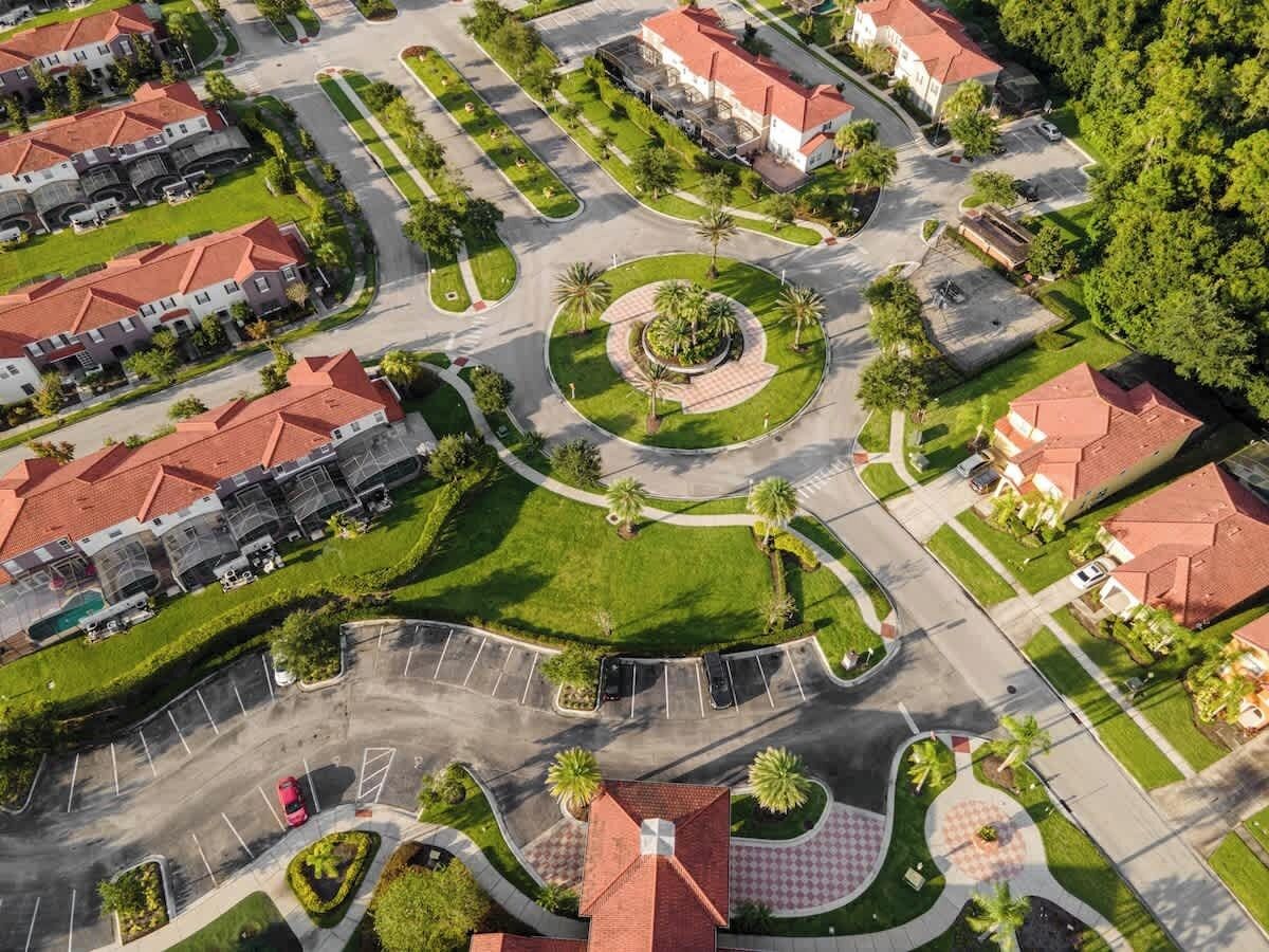An aerial view of a residential area with a central roundabout, surrounded by houses with red roofs, green lawns, and palm trees.