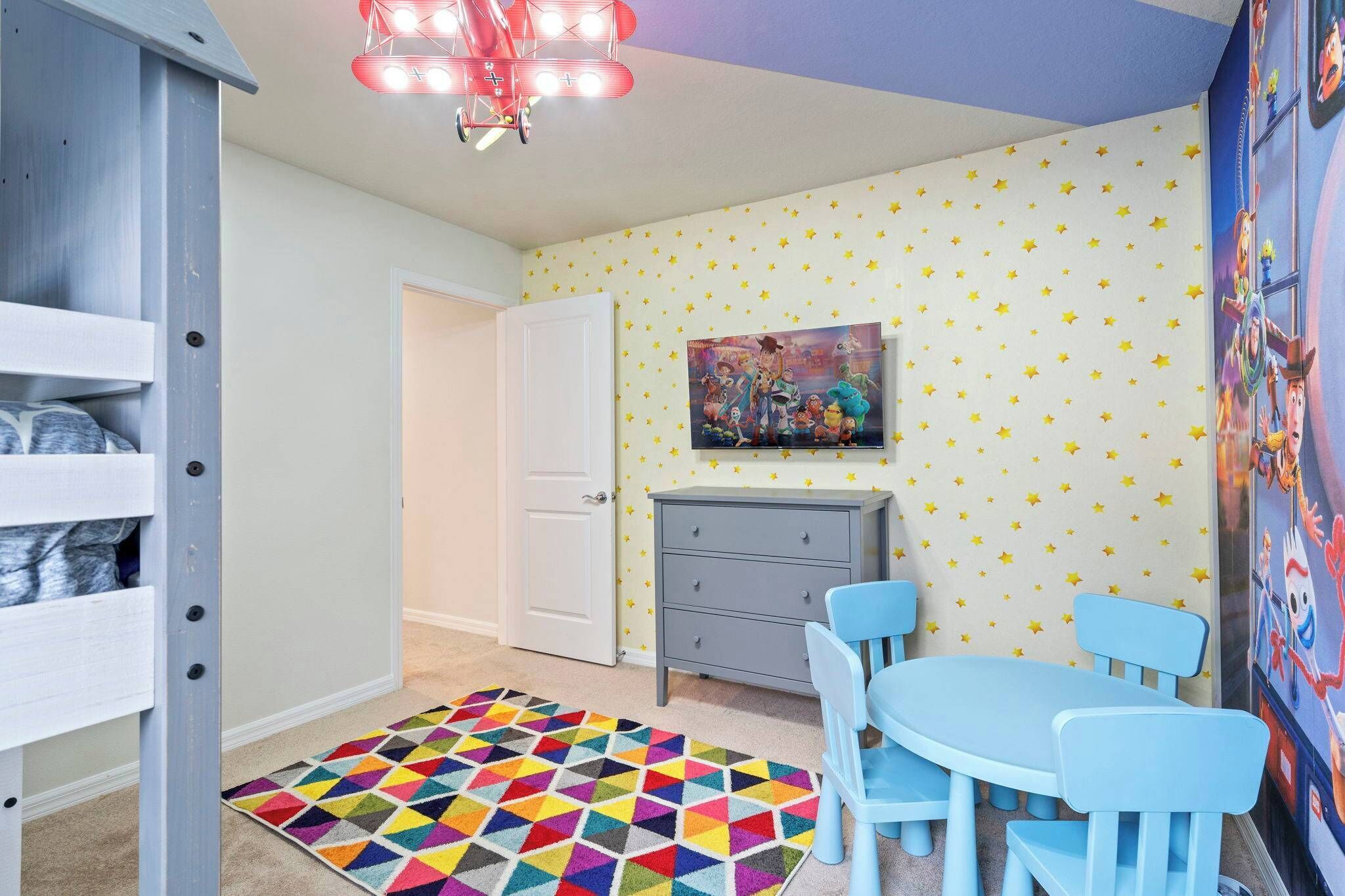 A child's room with a bunk bed, a dresser, a desk with chairs, a colorful rug, and star-patterned wallpaper.