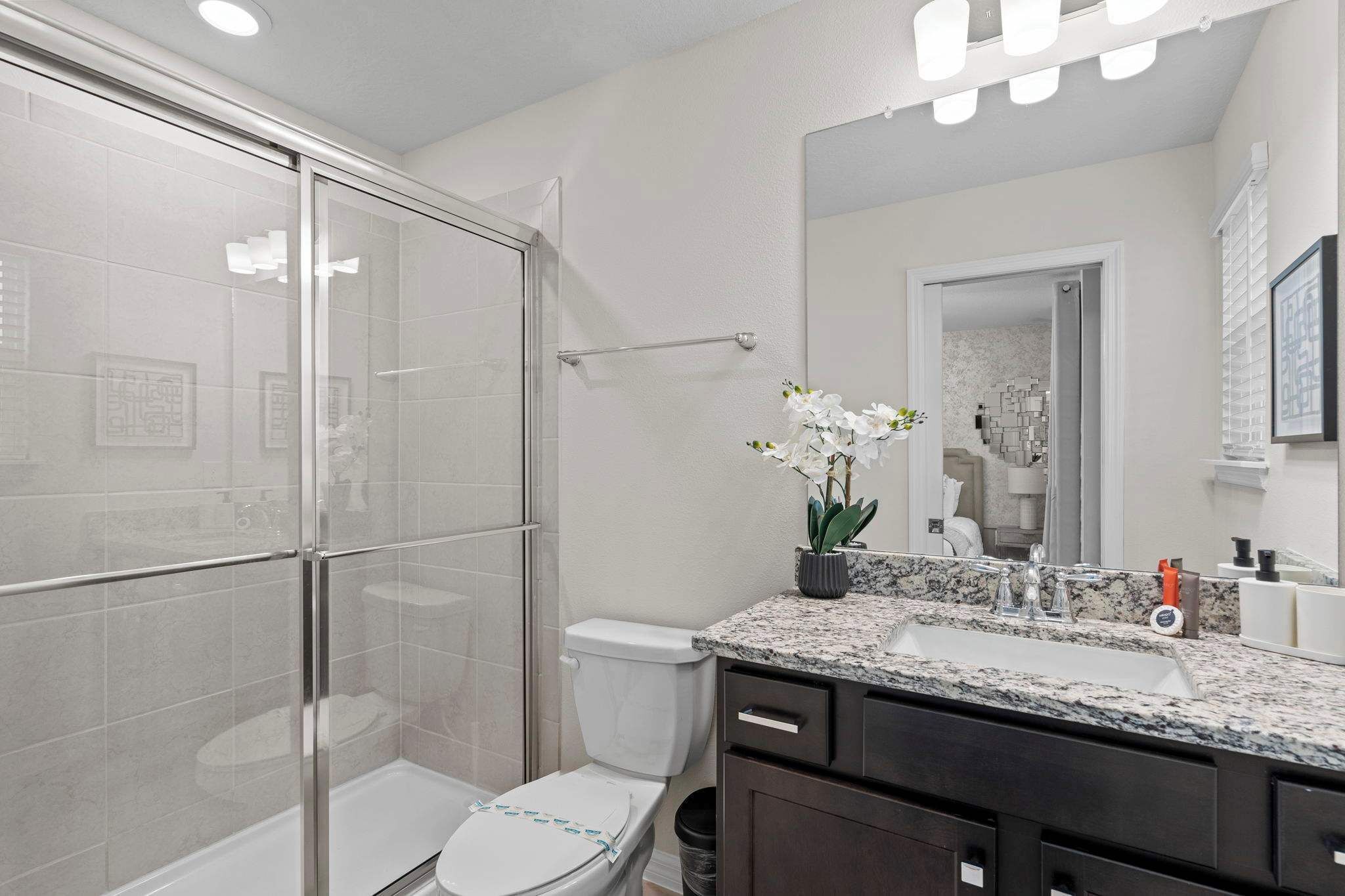 Un baño moderno con cabina de ducha de vidrio, inodoro, lavabo con encimera de granito y espejo sobre el lavabo.