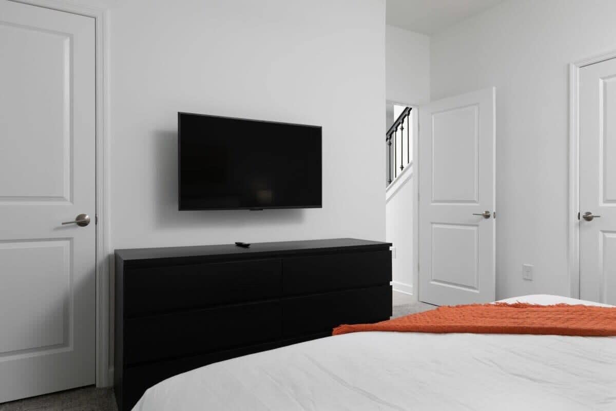 Une chambre à coucher avec un lit, une commode noire, un téléviseur fixé au mur et des portes blanches.