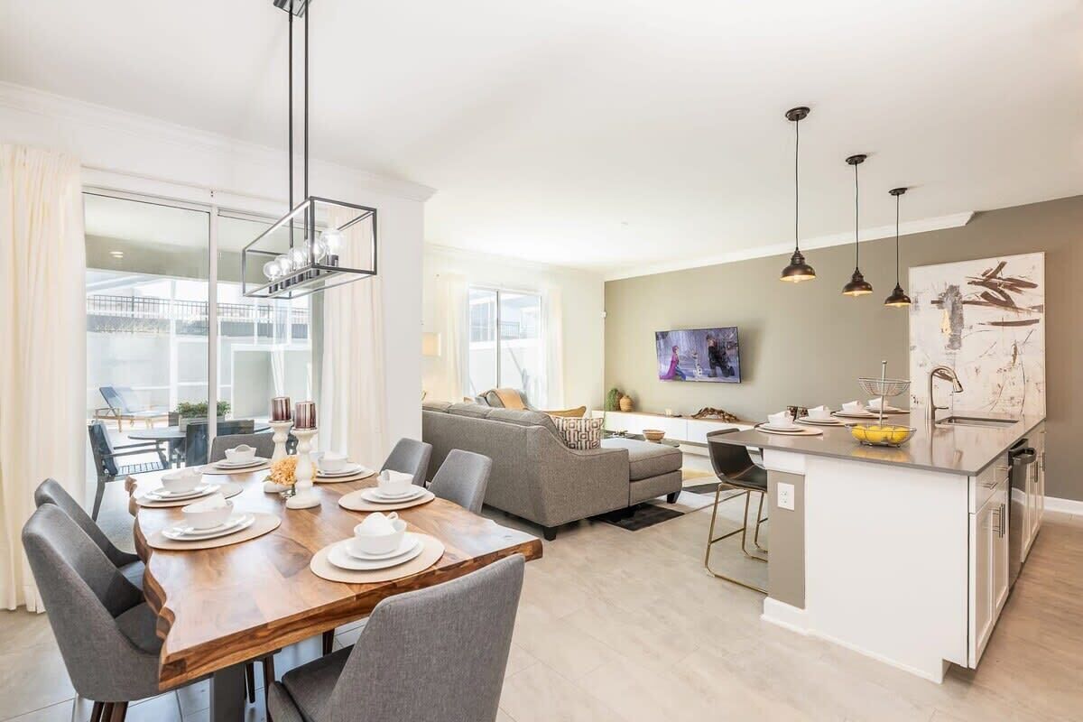 A modern living space with a dining area, a sofa, a kitchen island, pendant lighting, and a large window.